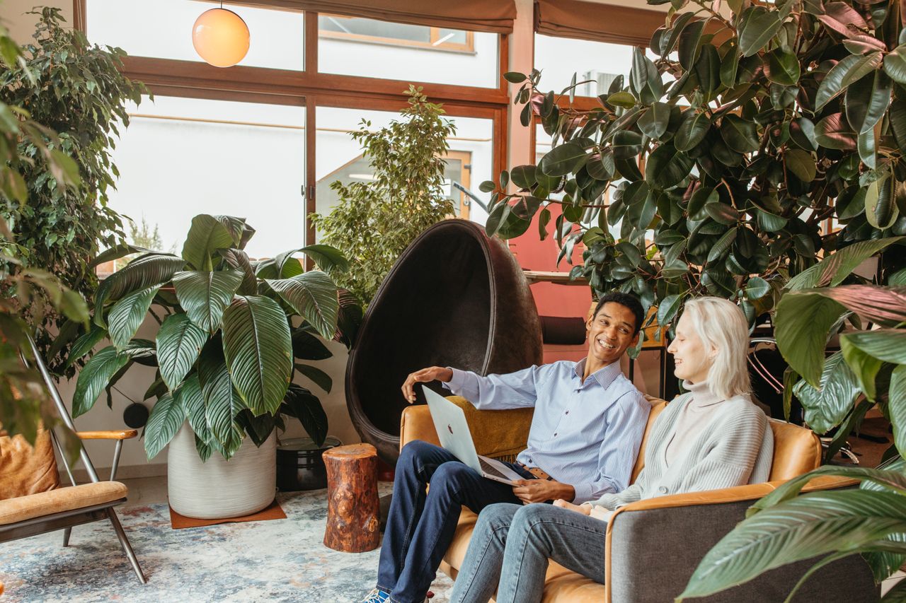 Diverse colleagues sit in a green-filled office lounge, discussing with smiles.